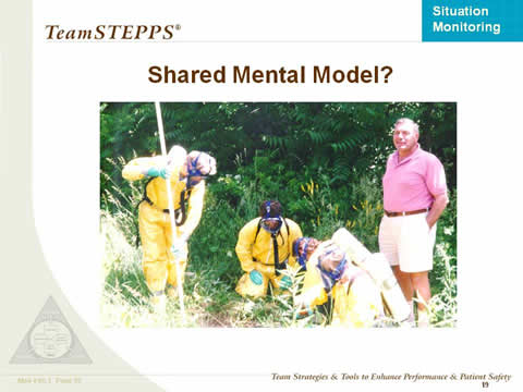 Shared Mental Model? Four people in a group are wearing hazard suits and masks and searching the ground. A fifth person in shorts and polo shirt stands by apparently unconcerned.